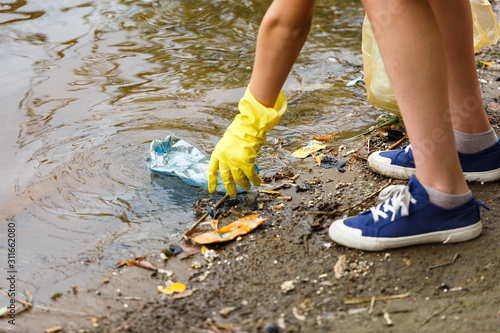 Volunteer boy in blue t shirt picks up dirty plastic bottles in park. Hands in yellow gloves collect garbage, pick up trash in bag. Eco activist cleaning up nature from plastic	