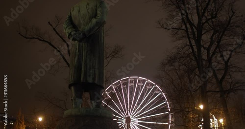 Medium full 4K shot with slow tilt reveal motion of Henrik Ibsen statue in front of National Theatre with illuminated Ferris wheel at local Christmas market in background, at night in Oslo Norway. photo