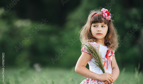 Little  beautiful and fragile Ukrainian girl on the background of nature with greens. Cute child portrait close-up with spikelet. Young child posing in colorful dress and bow. Natural photography.