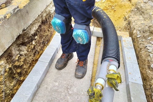 Worker in the knee pads. Black plastic thermally insulated pipe. Long water pipes with thermal insulation lie underground. View on assembled and isolated pipeline in trench.

