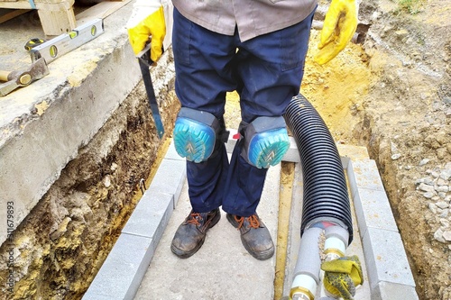 Worker in the knee pads. Black plastic thermally insulated pipe. Long water pipes with thermal insulation lie underground. View on assembled and isolated pipeline in trench.