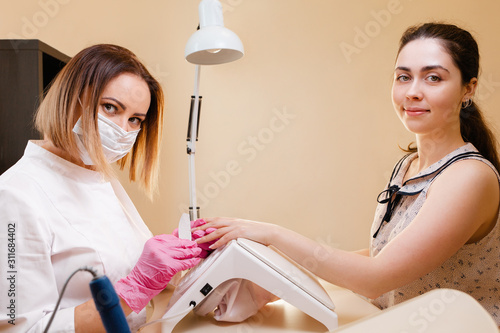 Medicine, cosmetology and manicure. A woman manicure master, in a mask and a white coat does a manicure to the client in the salon, filing her nails. Side view