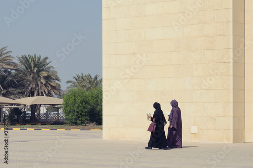 Two muslim woman wearing traditional long dress covered face walking and reading book holding water on big wall building background outdoor over sky, Bahrain, Unreconised person.