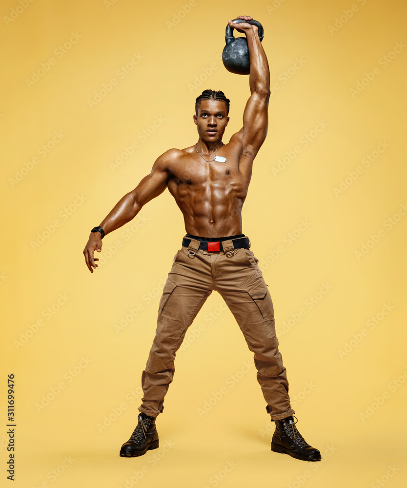 Young muscular guy training with kettlebell. Photo of handsome man with naked torso on yellow background. Strength and motivation. Full length