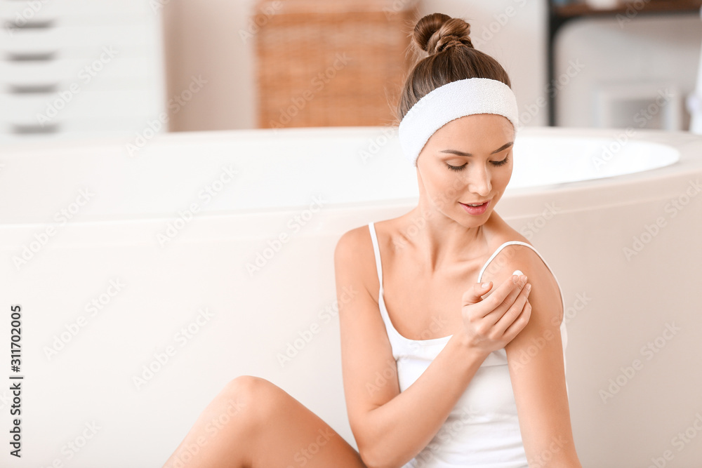 Beautiful young woman applying cream onto her skin in bathroom Beautiful young woman applying cream onto her skin in bathroom