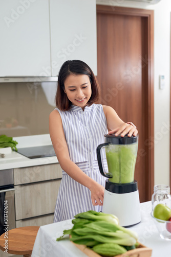 Pretty young Asian woman making smoothie in blender in the kitchen