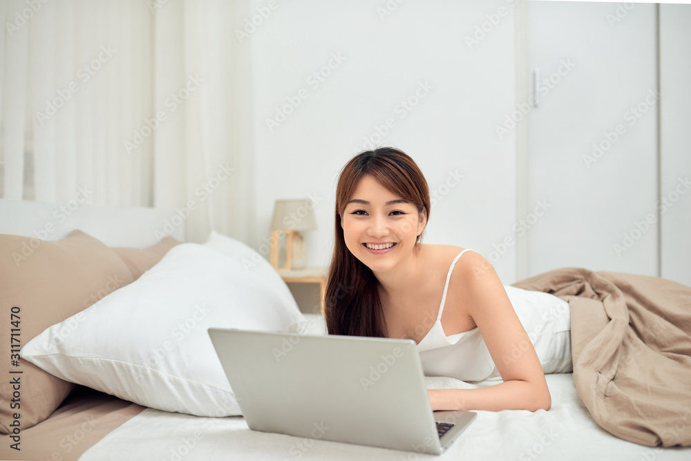 Smiling woman using a laptop while lying on her bed Smiling woman using a laptop while lying on her bed