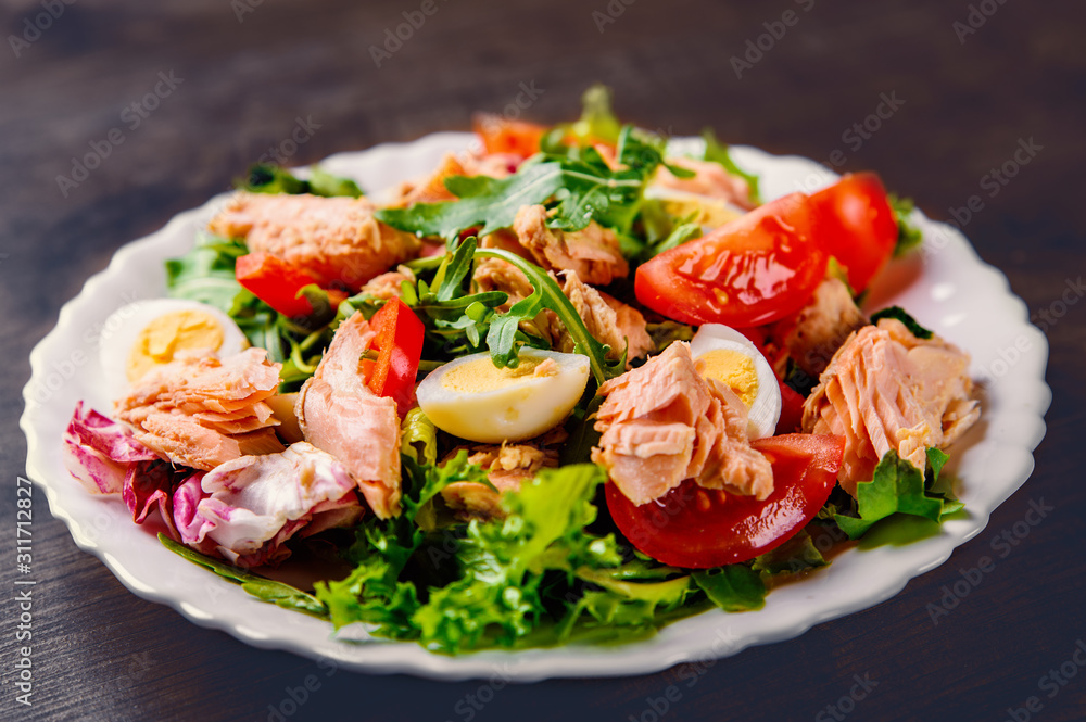 Fresh salad with fish, arugula, eggs,red pepper, lettuce, fresh sald leaves and tomato on a white plate on wooden table background
