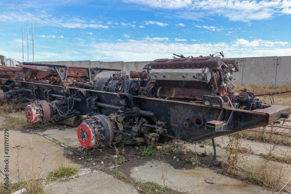 An abandoned rusty frame and part of the power plant and transmission of an old combat vehicle, standing without wheels, on blocks, in the open.