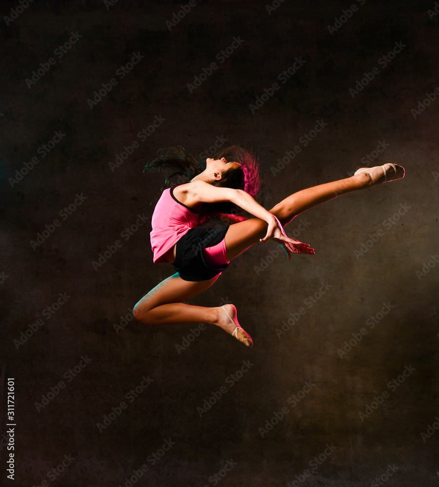 Young slim athletic girl gymnast dancer jumping up flying stretching doing gymnastic exercises in studio on dark wall