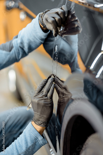 Worker trimming with cutter remains of a protective film, sticking it on a car body at the vehicle service, close-up. Concept of car body protection with special films