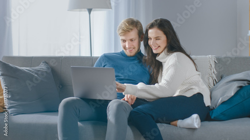 Cute Young Couple Use Laptop Computer, Have Fun, Laughs, while Sitting on the Couch in the Cozy Apartment. Couple Shopping on Internet, Using Social Media, Watching Videos and Streaming Content 