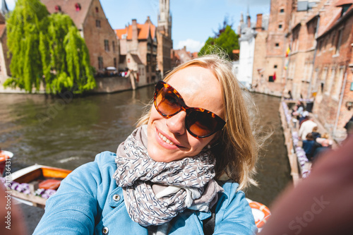 Beautiful young girl takes selfie photo on the background of the famous tourist destination with a canal in Bruges, Belgium