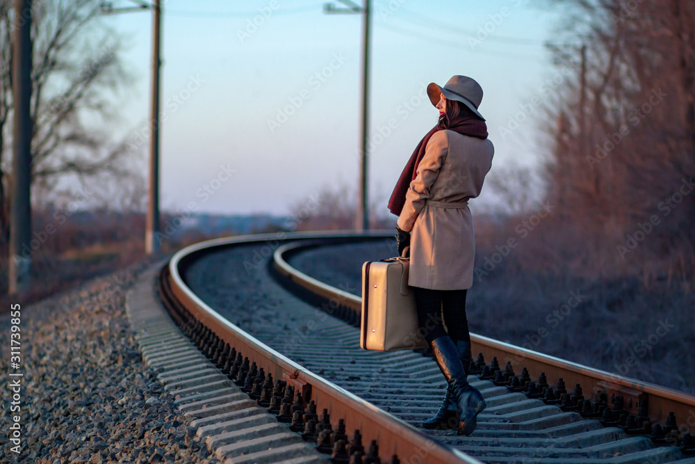 lonely young girl stands sideways with a beige suitcase in her hands on the railway at sunset