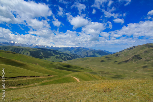 Road over green hills, mountains in snow and clouds in blue sky