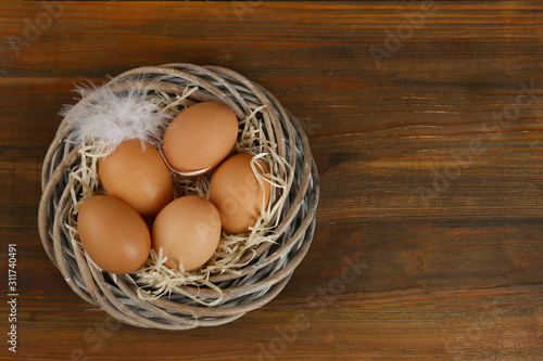 Raw chicken eggs and feather in wicker nest on wooden table, top view. Space for text
