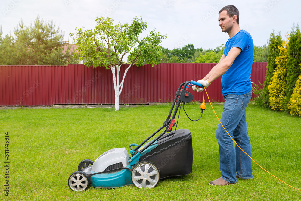 Fototapeta premium Worker with a lawn mower mows grass in the garden. Side view.