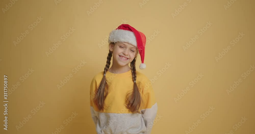 Little girl in santa hat smiling to the camera at yellow background slow motion Little girl in santa hat smiling to the camera at yellow background slow motion