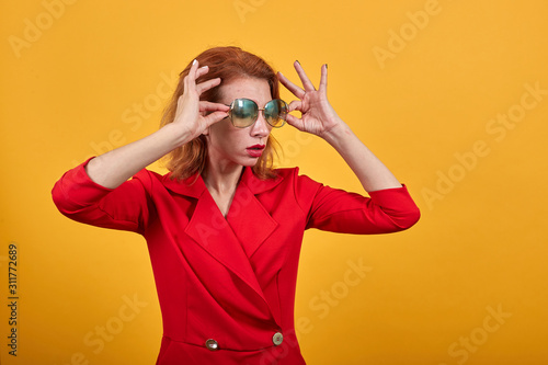 Charming caucasian young woman having fashion sunglasses, keeping hand on it, wearing fashion red jacket over isolated orange background. People lifestyle concept.