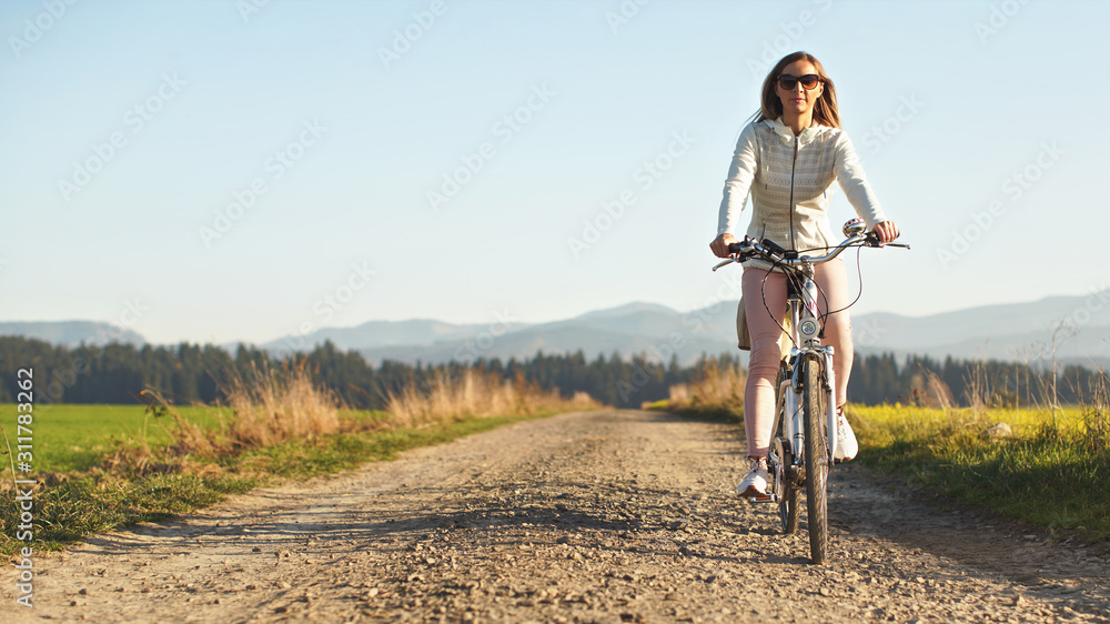Young woman in jacket and sunglasses rides a bicycle over country road, afternoon sun shines on blurred background, space for text left side Young woman in jacket and sunglasses rides a bicycle over country road, afternoon sun shines on blurred background, space for text left side
