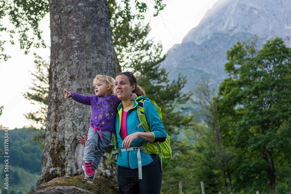 Mother supporting her daughter standing on tree trunk exploring the forest view in mountains.