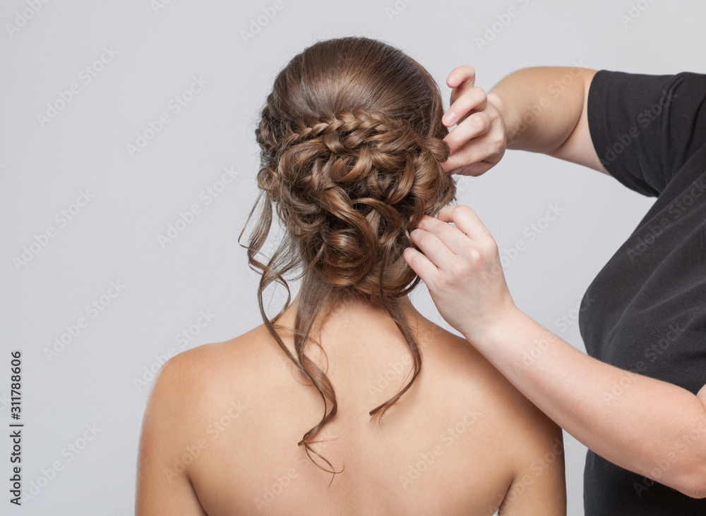 Naklejka premium Portrait of a beautiful sensual light brown haired woman with a wedding hairstyle in a beauty salon. The hairdresser does the hairstyle. Wedding hairstyle.