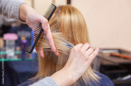 The hairdresser does a haircut with scissors of hair to a young girl, a blonde in a beauty salon. Professional hair care.