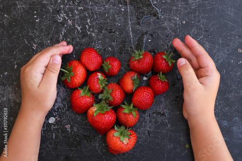 Red juicy strawberries on a black table.  The berries are stacked in the shape of a heart.  Hands of a child on the table.