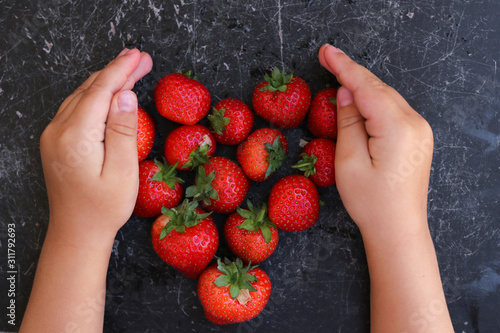 Red juicy strawberries on a black table.  The berries are stacked in the shape of a heart.  Hands of a child on the table.