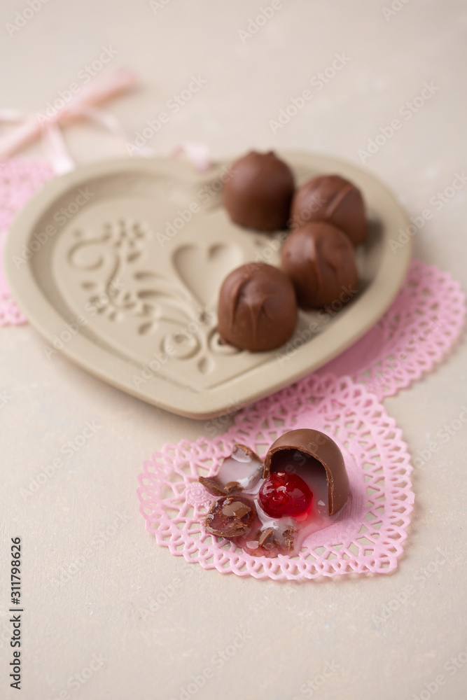 Chocolate Covered Cherries displayed in a heart shaped cookie mold. One isolated in front on pink heart-shaped doilies. Beige background with room for text. Chocolate Covered Cherries displayed in a heart shaped cookie mold. One isolated in front on pink heart-shaped doilies. Beige background with room for text.