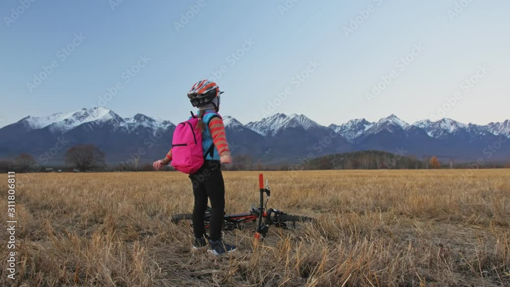 One caucasian children walk with bike in wheat field. Girl walking black orange cycle on background of beautiful snowy mountains. Biker motion ride with backpack and helmet. Mountain bike hardtail. One caucasian children walk with bike in wheat field. Girl walking black orange cycle on background of beautiful snowy mountains. Biker motion ride with backpack and helmet. Mountain bike hardtail.