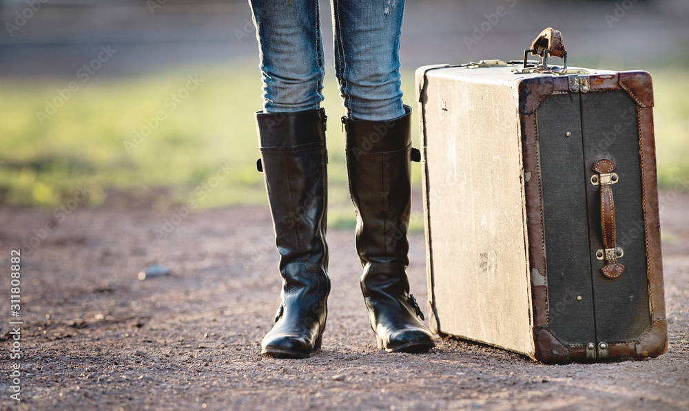 Person with tall black boots standing next to a vintage suitcase on a gravel road Person with tall black boots standing next to a vintage suitcase on a gravel road