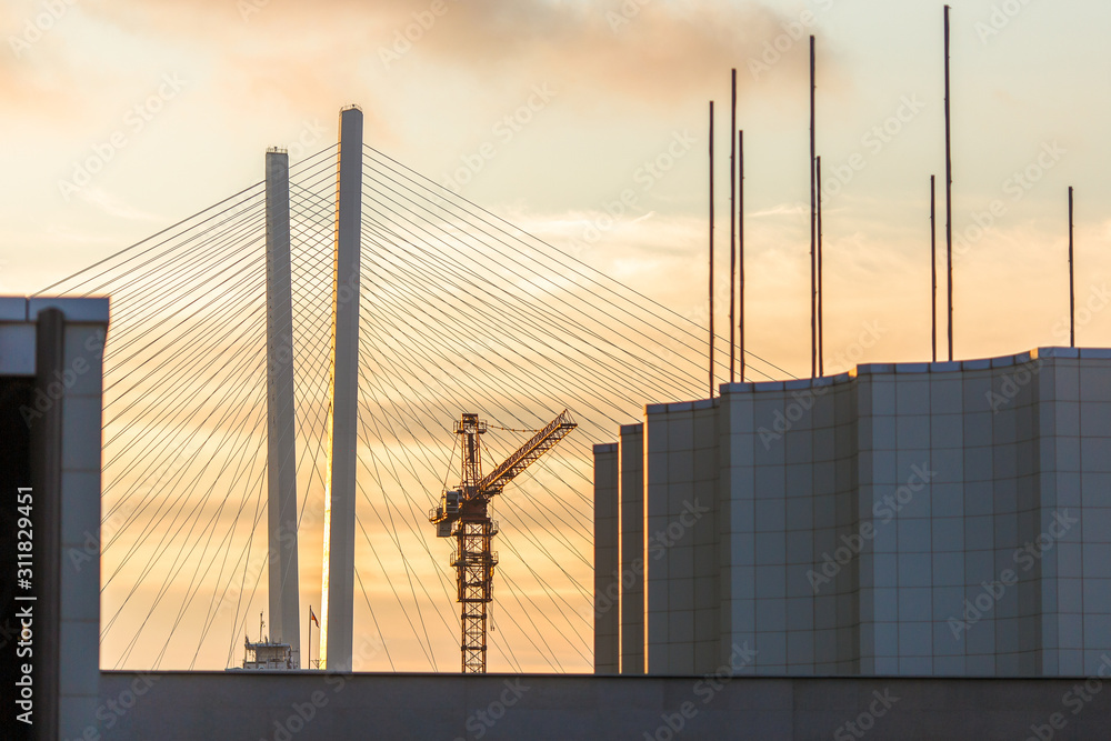 Construction crane on the background of the Golden Bridge in Vladivostok. Tower crane on a background of red golden dawn. Construction crane on the background of the Golden Bridge in Vladivostok. Tower crane on a background of red golden dawn.