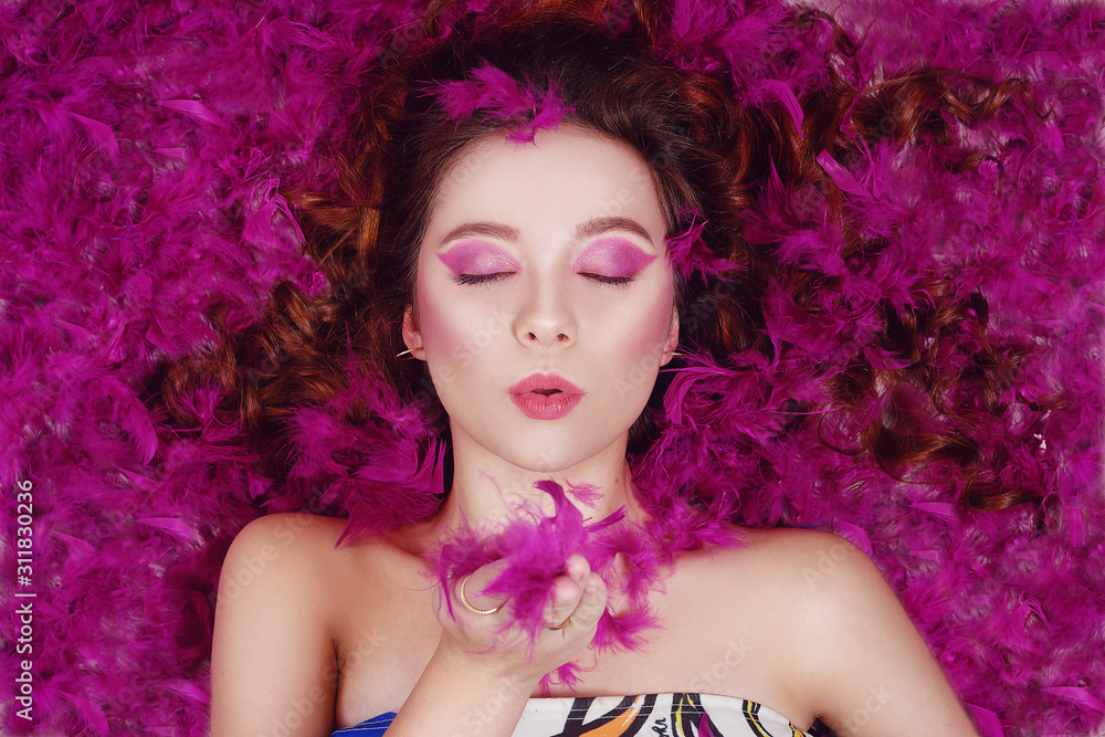 A beautiful girl with professional purple make-up sends an air kiss to the camera. The model lies in purple feathers. Fashion photo on a pink background. A beautiful girl with professional purple make-up sends an air kiss to the camera. The model lies in purple feathers. Fashion photo on a pink background.