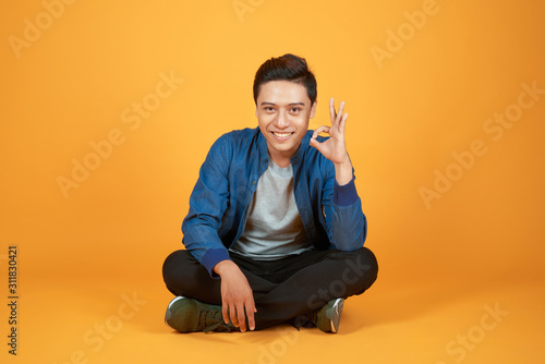 Appealing young Asian man sitting with his legs crossed and showing okay sign. Isolated on orange background © makistock