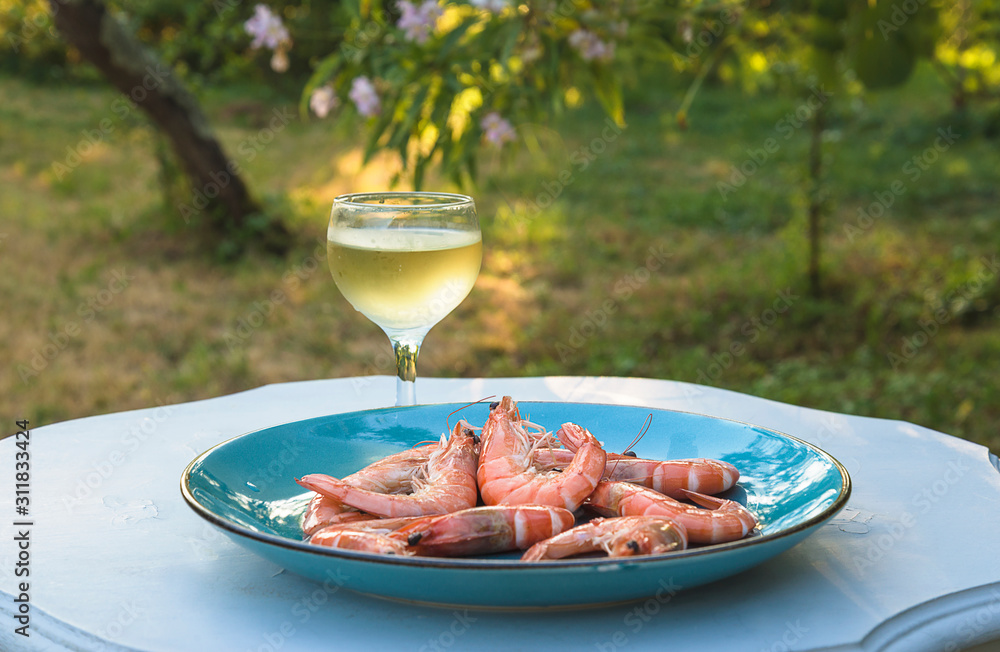 Pink prawns or shrimps on the blue plate and glass of cold white wine on the white table in the blooming garden on sunny summer day