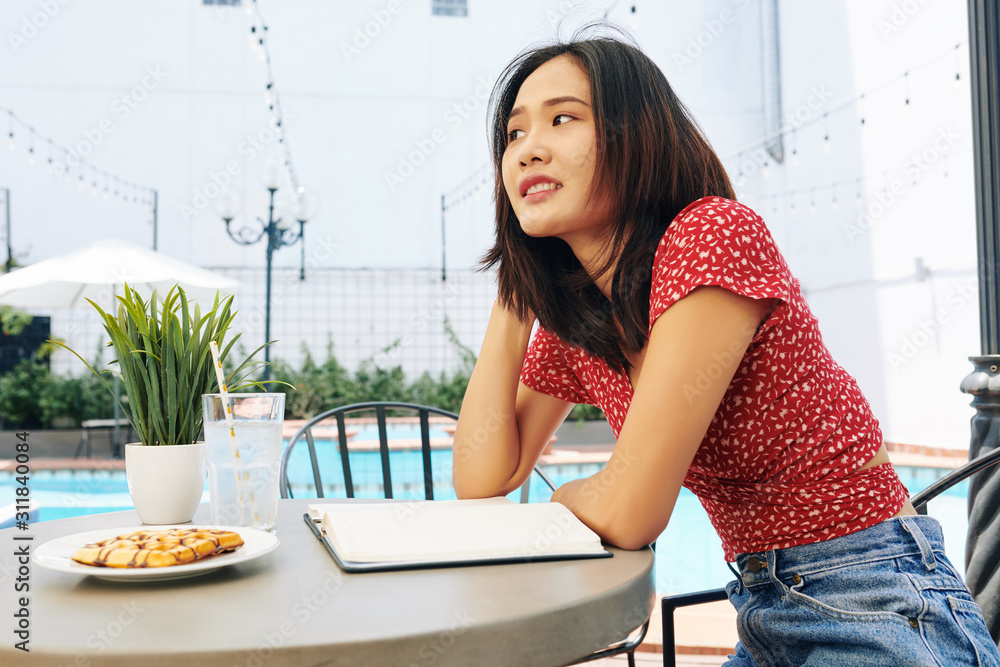 Portrait of young pensive Vietnamese woman sitting at cafe table with opened planner and breakfast Portrait of young pensive Vietnamese woman sitting at cafe table with opened planner and breakfast
