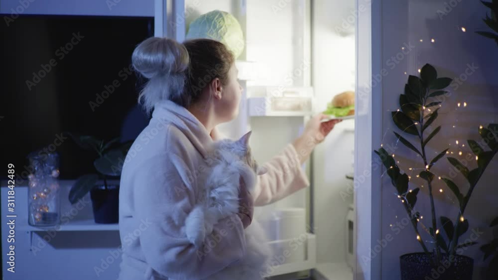 eating disorder, hungry woman with a cat in her arms opens a refrigerator at night and inhales the aroma of a hamburger then go away in room with a plate eating disorder, hungry woman with a cat in her arms opens a refrigerator at night and inhales the aroma of a hamburger then go away in room with a plate