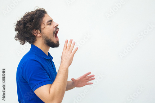 Side of excited serious guy shouting loud at copy space. Handsome bearded young man in blue casual t-shirt posing isolated over white background. Announcement concept