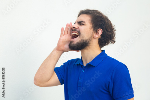 Excited guy sharing important news, shouting loud with hand at open mouth. Handsome bearded young man in blue casual t-shirt posing isolated over white background. Advertising concept © Mangostar