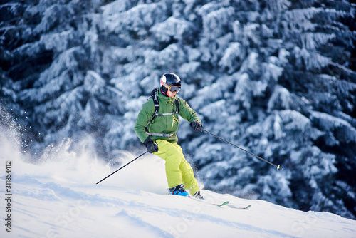 Proficient skier coming down along wooded hillside using professional ski equipment and making deep snow powder. Winter outdoors activities concept. Picturesque forest scenery on background. Side view