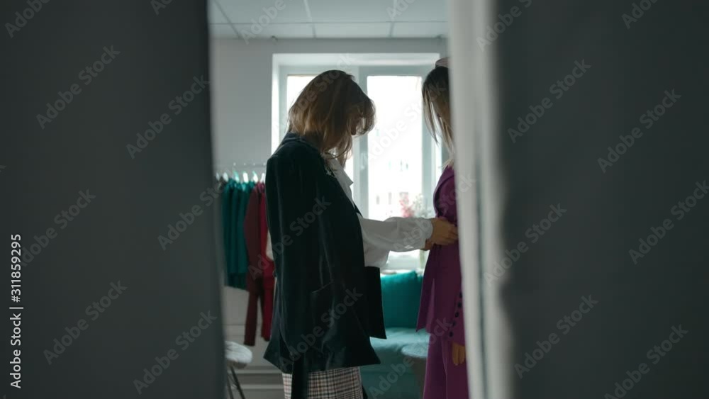 Side view of young woman coming closer to friend and adjusting stylish jacket while visiting clothes shop together. Side view of young woman coming closer to friend and adjusting stylish jacket while visiting clothes shop together.
