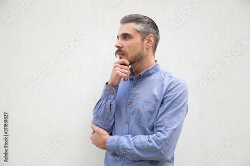 Pensive man touching chin and looking away at copy space. Grey haired young man in blue casual shirt posing isolated over white background. Advertising concept