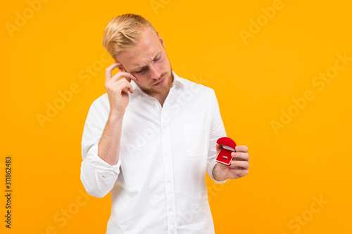 Young beautiful man holds an egagement ring for his girlfriend and call the phone isolated on yellow background photo