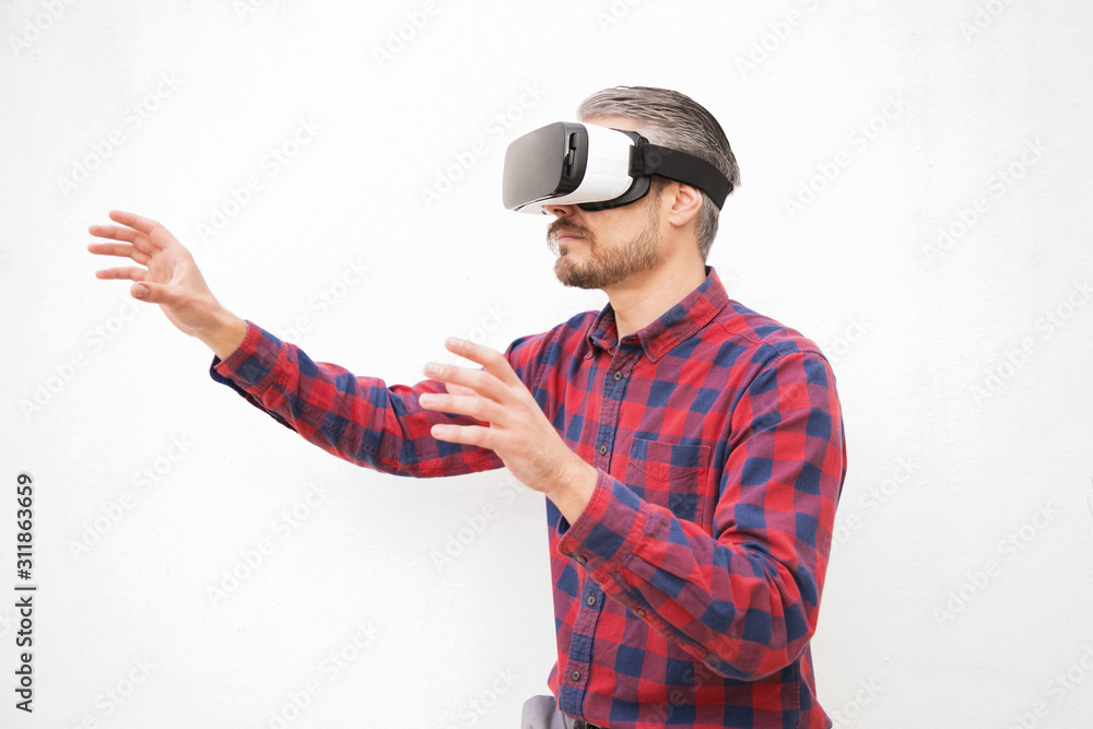 Man in VR headset touching air. Serious bearded man in checkered shirt moving with hands and using VR headset on grey background. Technology concept Man in VR headset touching air. Serious bearded man in checkered shirt moving with hands and using VR headset on grey background. Technology concept