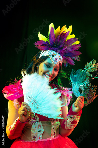 Beautiful young woman in carnival mask and stylish masquerade costume with feathers fan in colorful lights and glow on black background. Christmas, New Year, celebration. Festive time, dance, party.