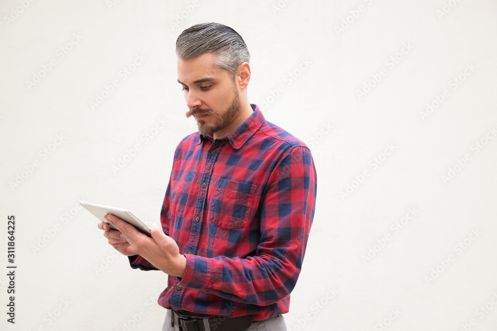 Concentrated man using tablet computer. Serious handsome man in checkered shirt standing and using digital tablet on grey background. Wireless technology concept Concentrated man using tablet computer. Serious handsome man in checkered shirt standing and using digital tablet on grey background. Wireless technology concept