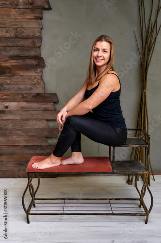 Portrait of a pretty fitness blonde girl in a tracksuit on an alternative background in vintage interior. Cute looks and smiles at the camera, sitting on a stand straight. Loft style concept.