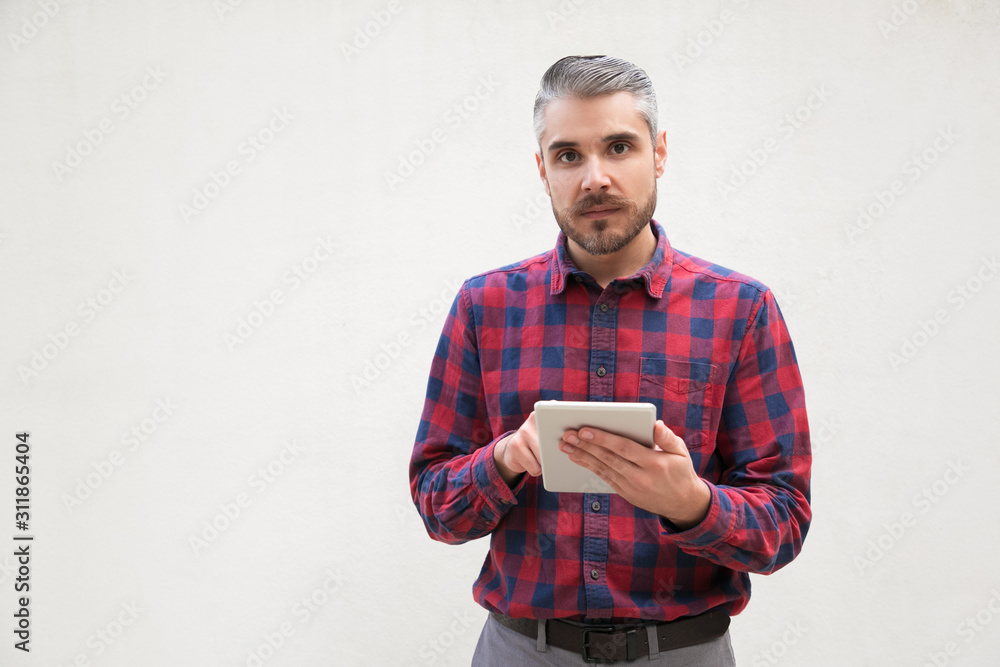 Serious man with digital tablet looking at camera. Portrait of handsome bearded man in checkered shirt using tablet pc on grey background. Wireless technology concept Serious man with digital tablet looking at camera. Portrait of handsome bearded man in checkered shirt using tablet pc on grey background. Wireless technology concept