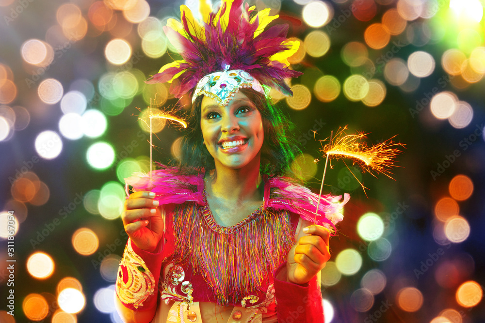 Fototapeta premium Beautiful young woman in carnival mask and stylish masquerade costume with feathers and sparklers in colorful bokeh on black background. Christmas, New Year, celebration. Festive time, dance, party.